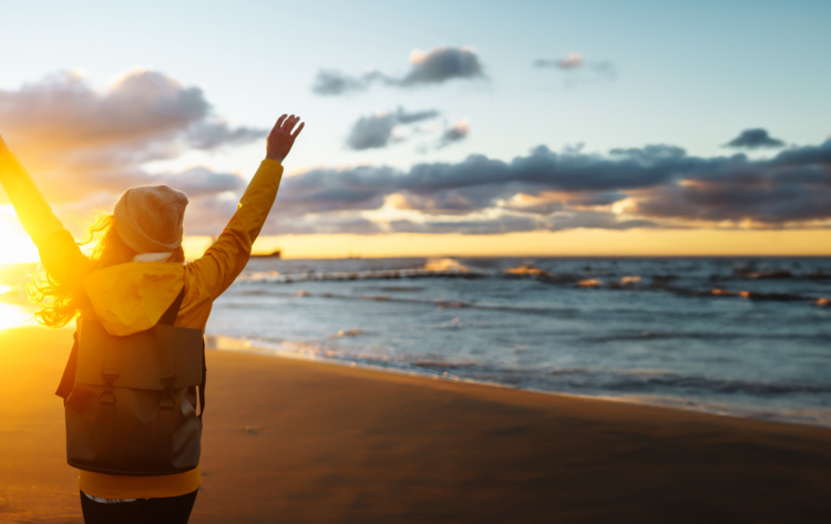 Eine Frau mit erhobenen Armen steht am Strand in Olpenitz beim Sonnenuntergang.